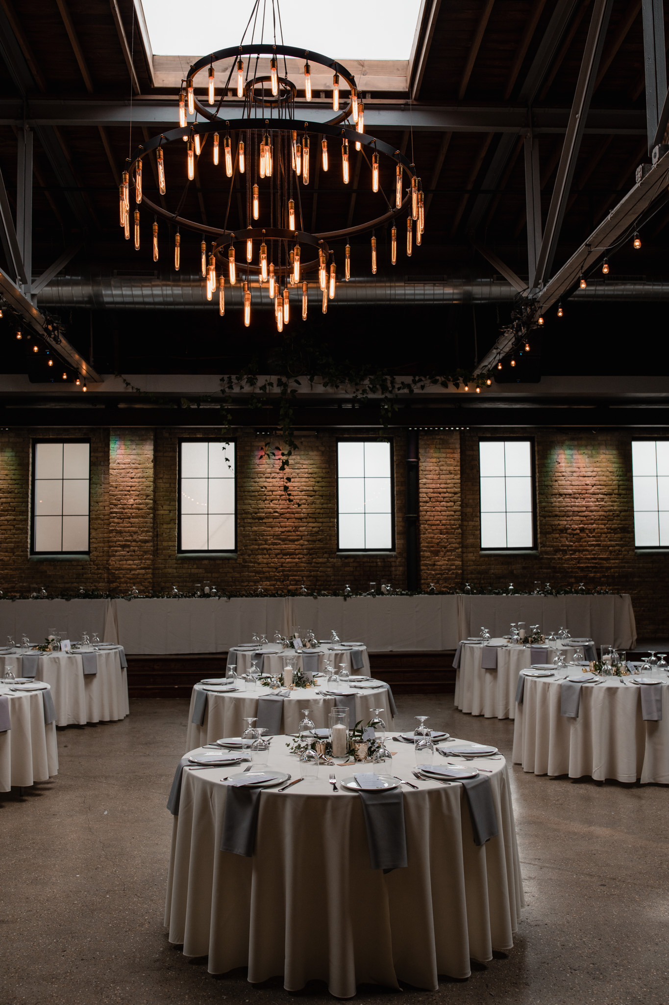 Inside of Milwaukee wedding venue Ivy House - Large windows, cream city brick, industrial chandelier, industrial ceiling, real plants. Lavender, white and green timeless table setting with round tables. Modern wedding photographer