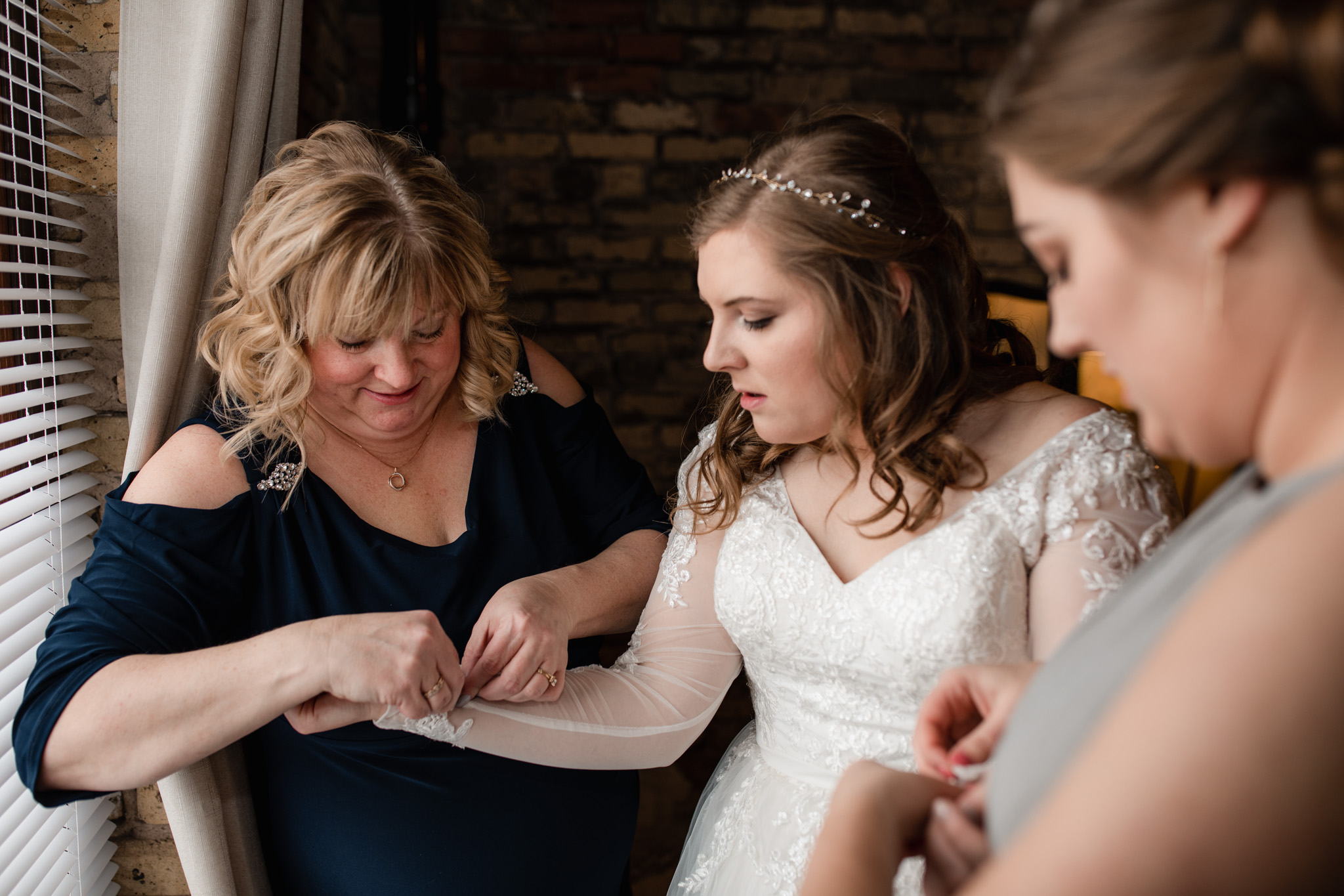 Mother and sister of the bride helping her get into her long sleeve lace dress in the getting ready room at Ivy House Milwaukee