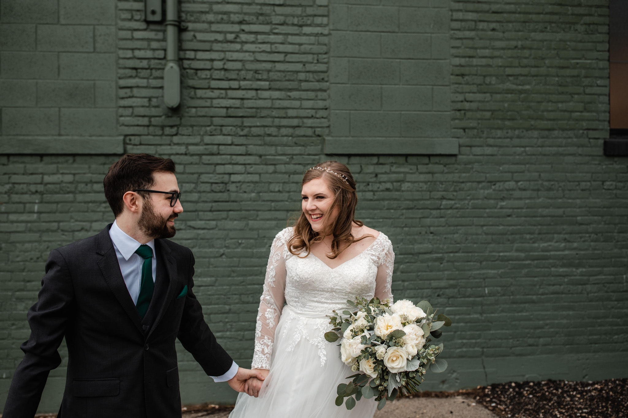 Couple laughing on their wedding day as they walk hand in hand. in front of a green wall