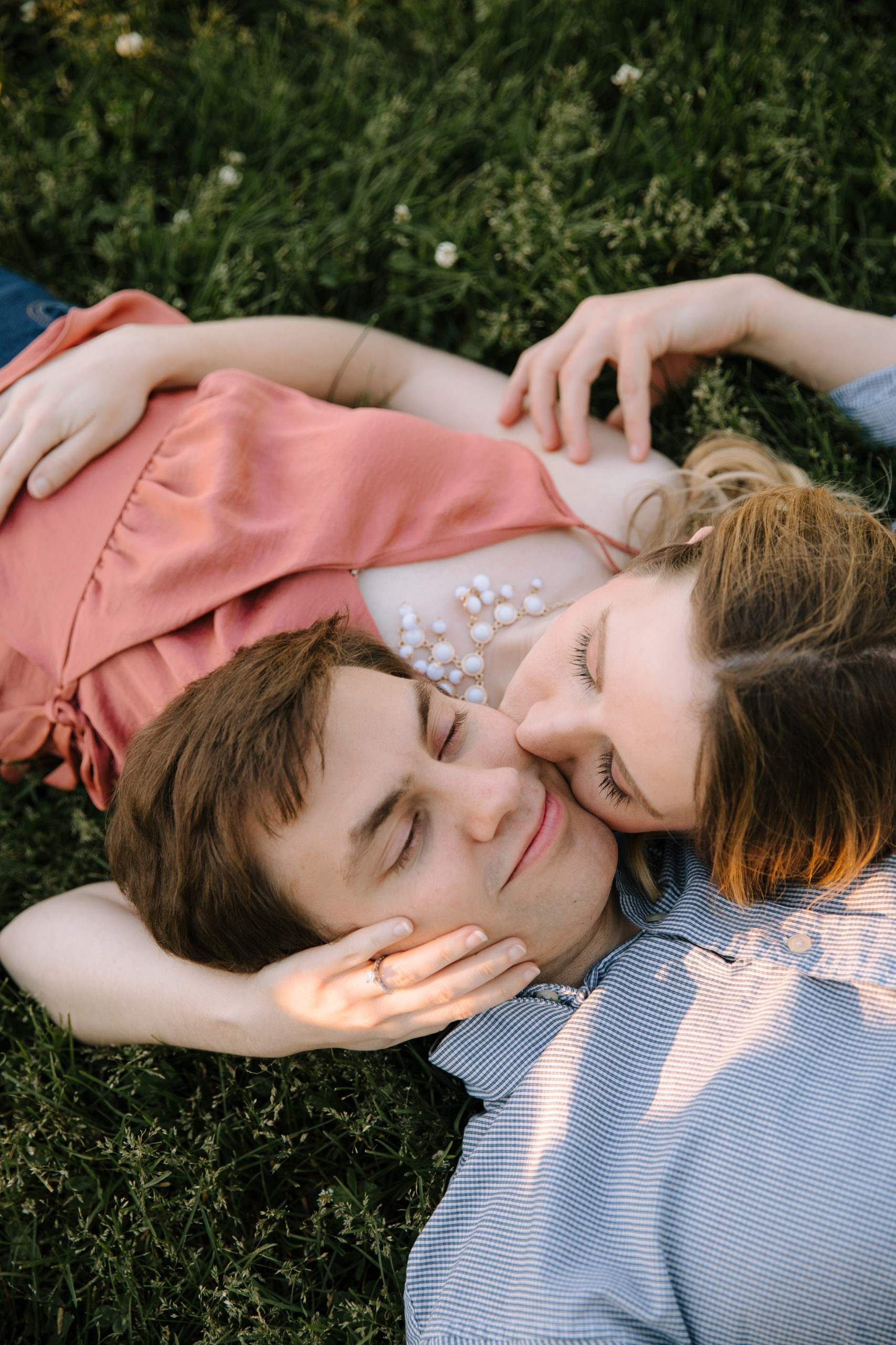 Couple laying in grass, head on each other's shoulders, girl kissing guy on the cheek with her ring hand on his other cheek. Guy is reaching back to touch girl's shoulder.