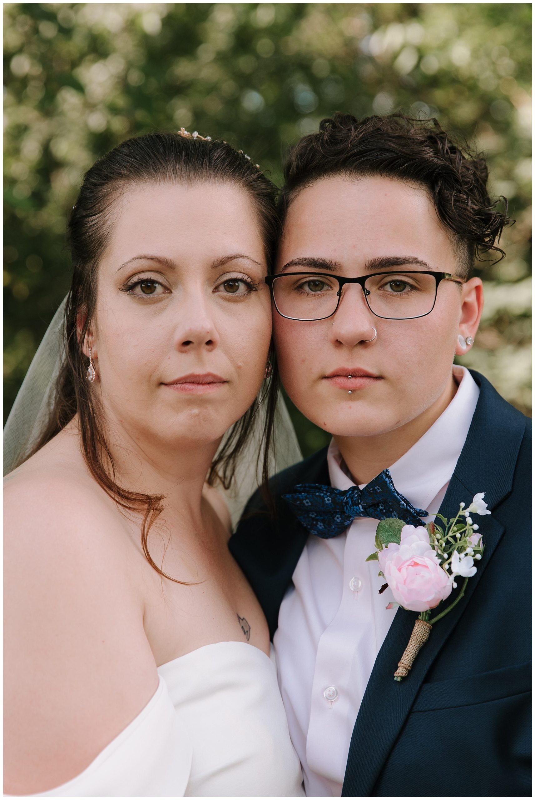 Two brides cheek to cheek looking at the camera. One bride is in a custom blue suit with a floral bowtie and the other bride is wearing a strapless dress with a veil and a curl sits over her shoulder.