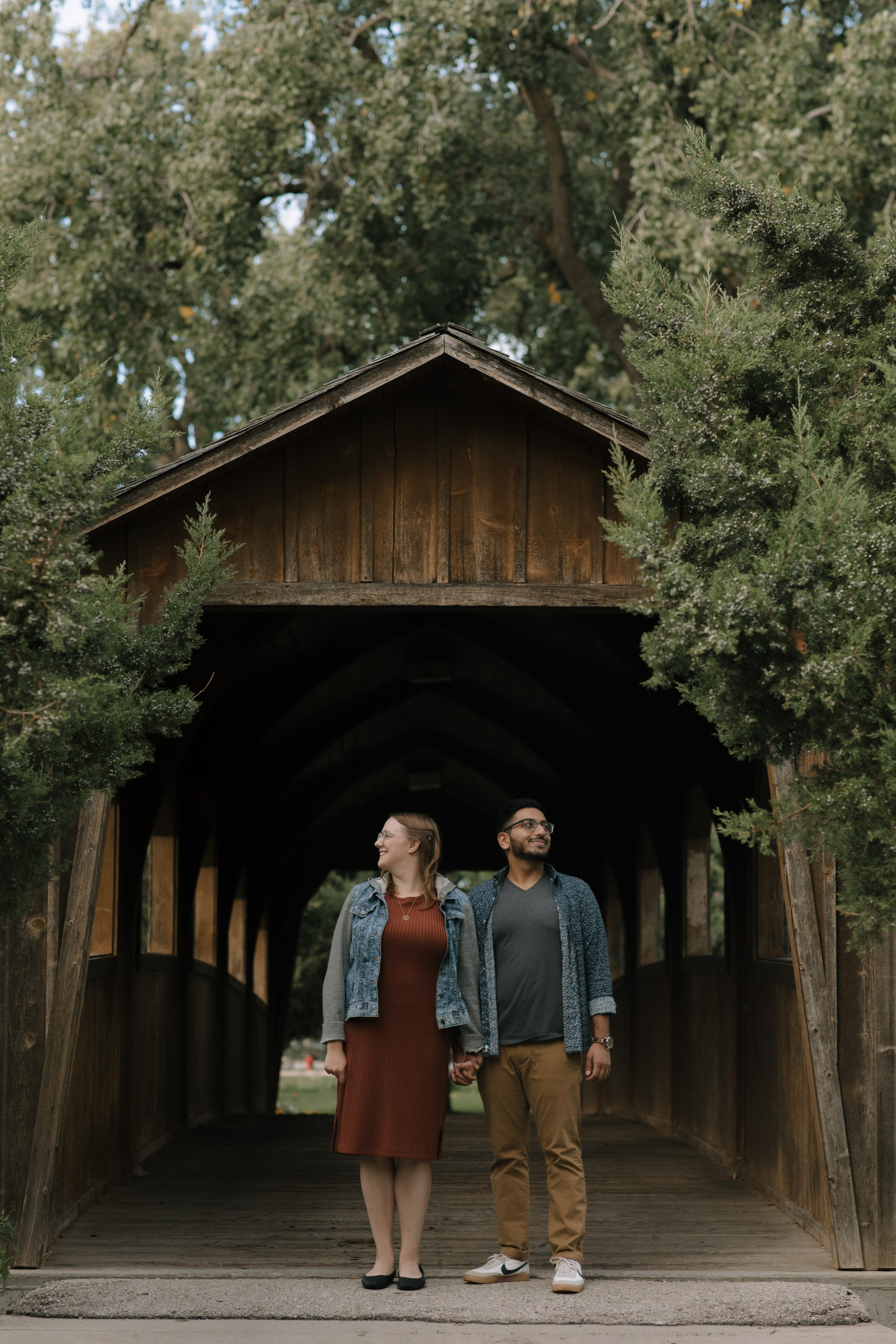 Couple wearing trendy outfits under a covered bridge for their engagement pictures in Fond Du Lac Wisconsin
