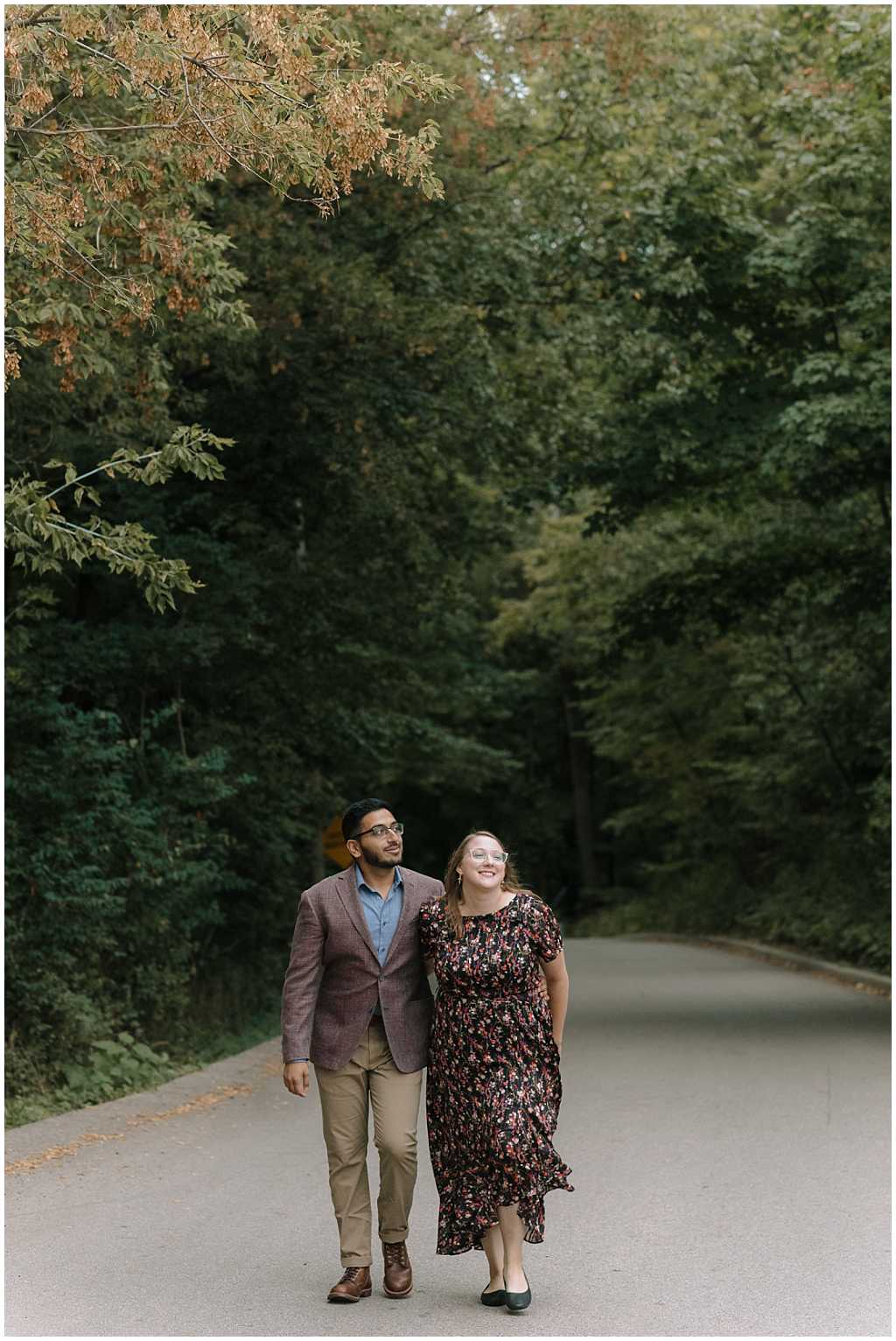 Couple walking along road in a forest. Man wearing purple suit jacket and khaki pants. Woman wearing black and pink flowy floral dress.