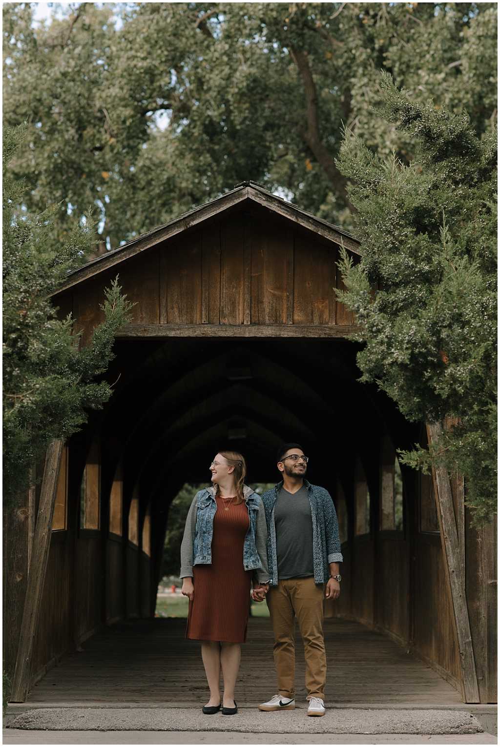 Couple standing side by side holding hands under covered bridge in Fond Du Lac Wisconsin. Woman wears ribbed knit orange dress with denim jacket. Man wears khaki pants with a gray v-neck and navy floral button up shirt for engagement pictures.