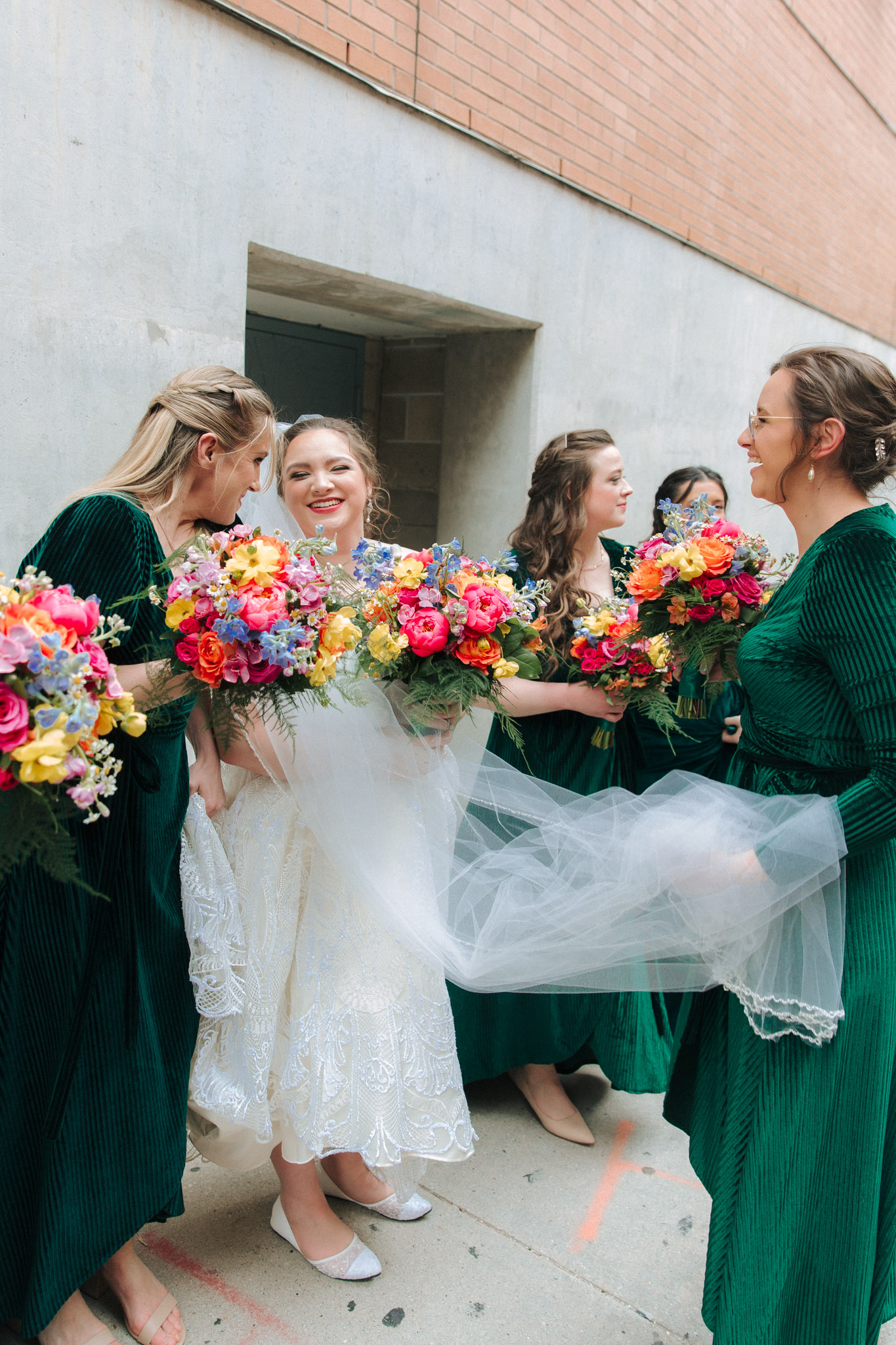 Candid of bridesmaids giggling with bride in emerald dresses with bright flowers outside of Pabst Theatre for wedding pictures