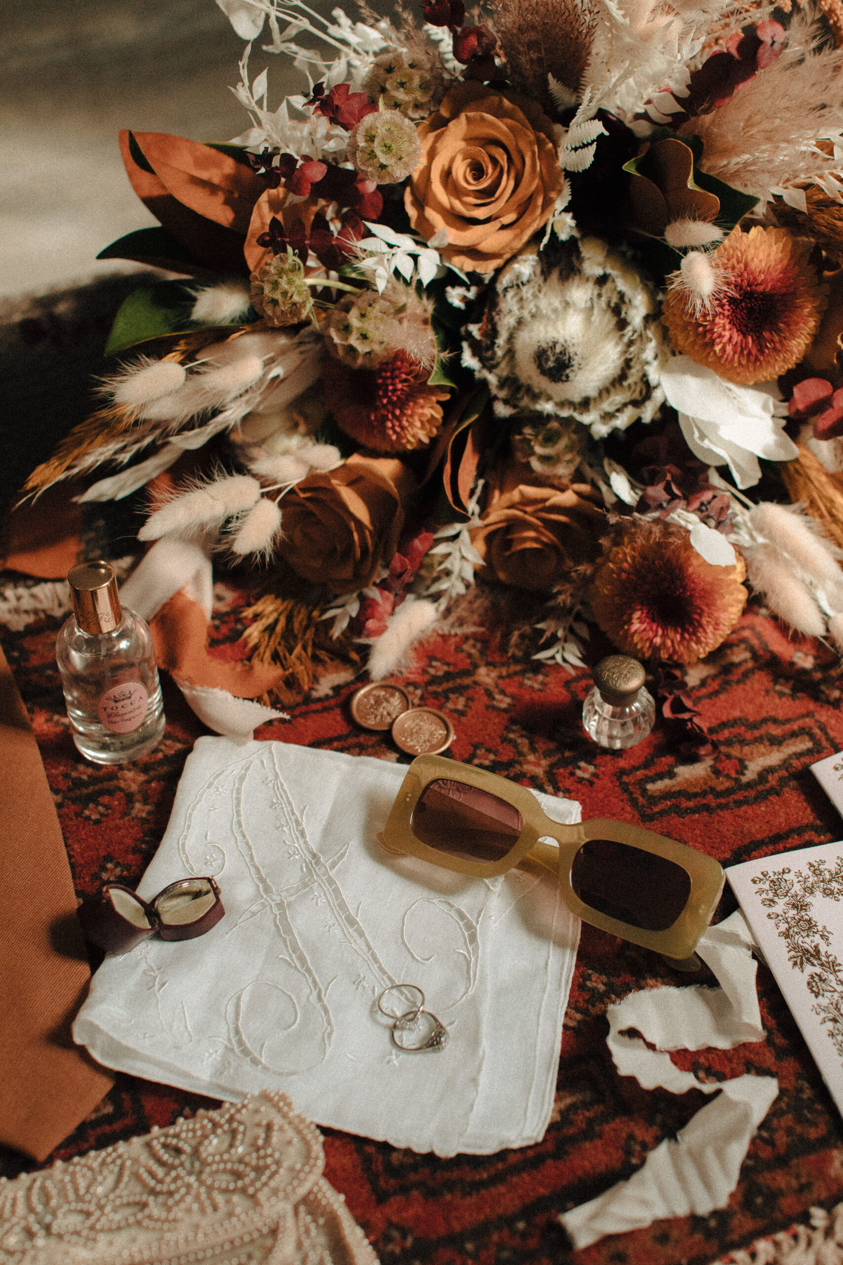 Vintage wedding flatlay details including men's vintage wedding band, monogrammed hankie, retro green sunglasses, boho bouquet, and beaded vintage clutch