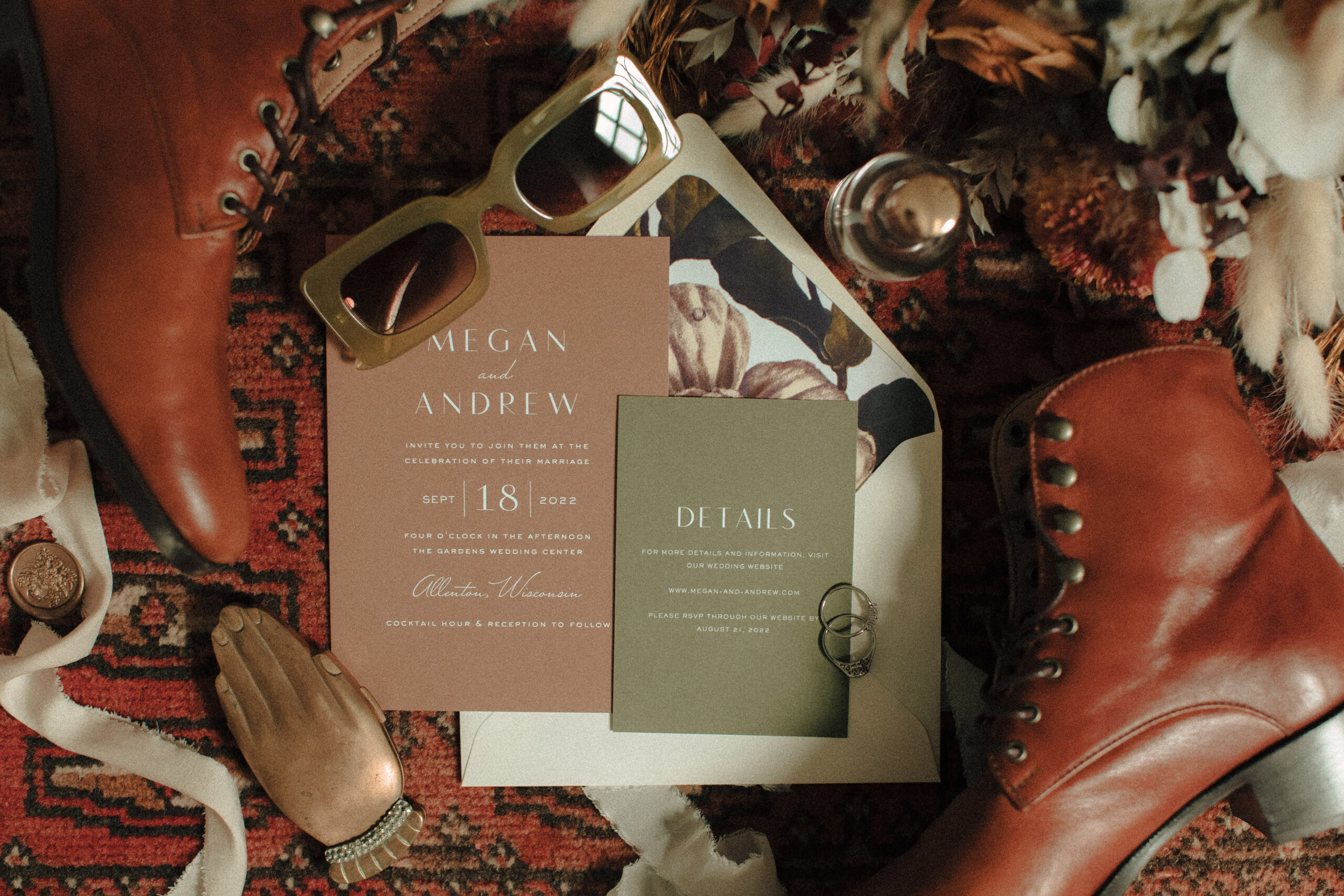 Vintage wedding flatlay details including women's boots, red rug, tan invitation and green detail card, vintage perfume bottle, wax seals, green retro sunglasses, and vintage engagement rings with boho flowers