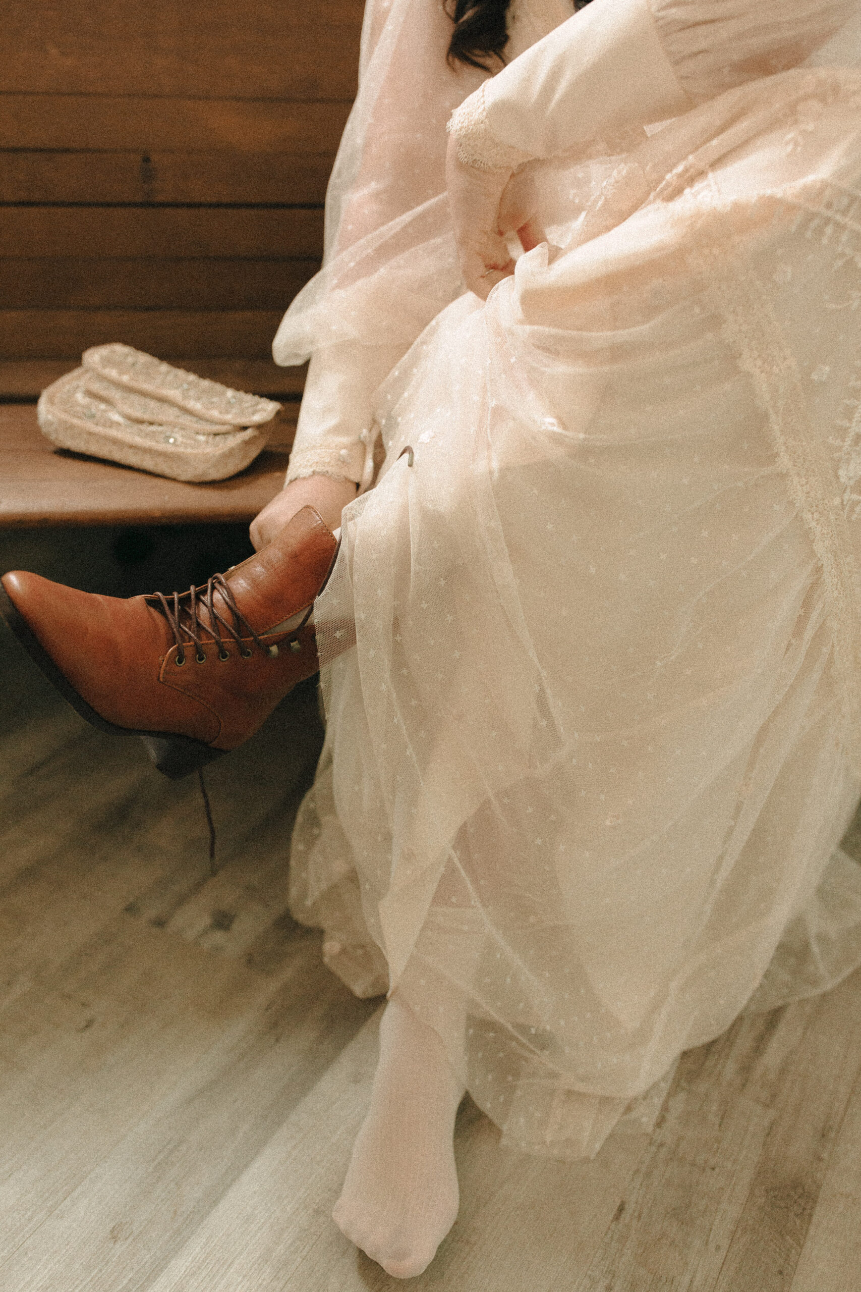 Bride wearing vintage wedding boots and creamy flowy gown with a vintage beaded clutch putting on her shoes