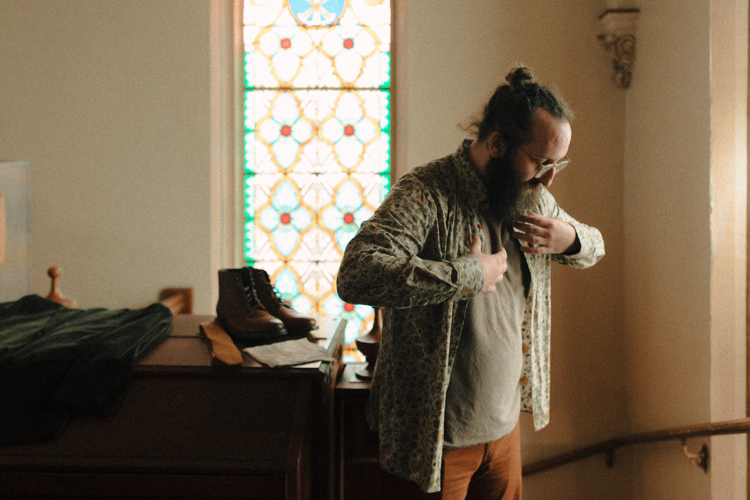 Groom with a beard and his hair in a bun getting ready in front of a stained glass window wearing a green paisley shirt and tan chinos