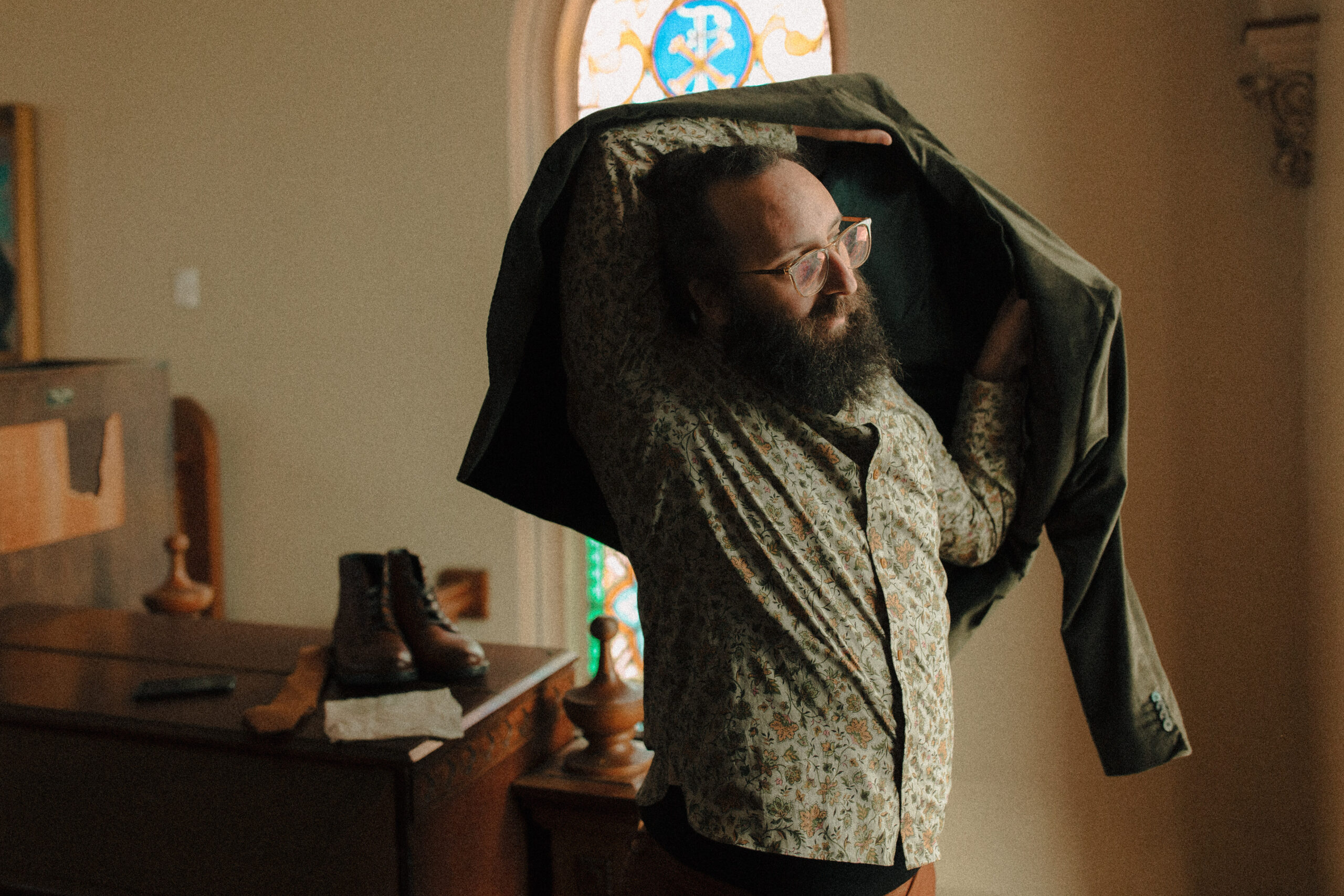 Groom with a beard and his hair in a bun getting ready in front of a stained glass window wearing a green paisley shirt and tan chinos