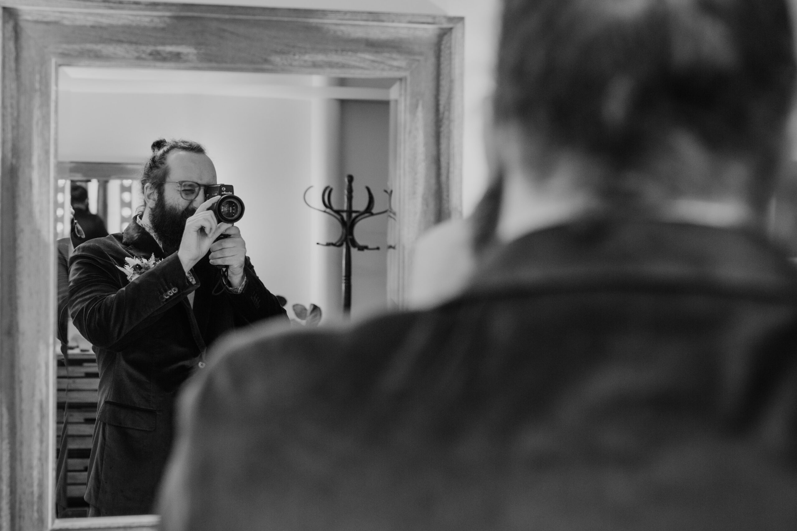 black and white photo of a groom using a super 8 camera on his wedding day to film himself in a mirror
