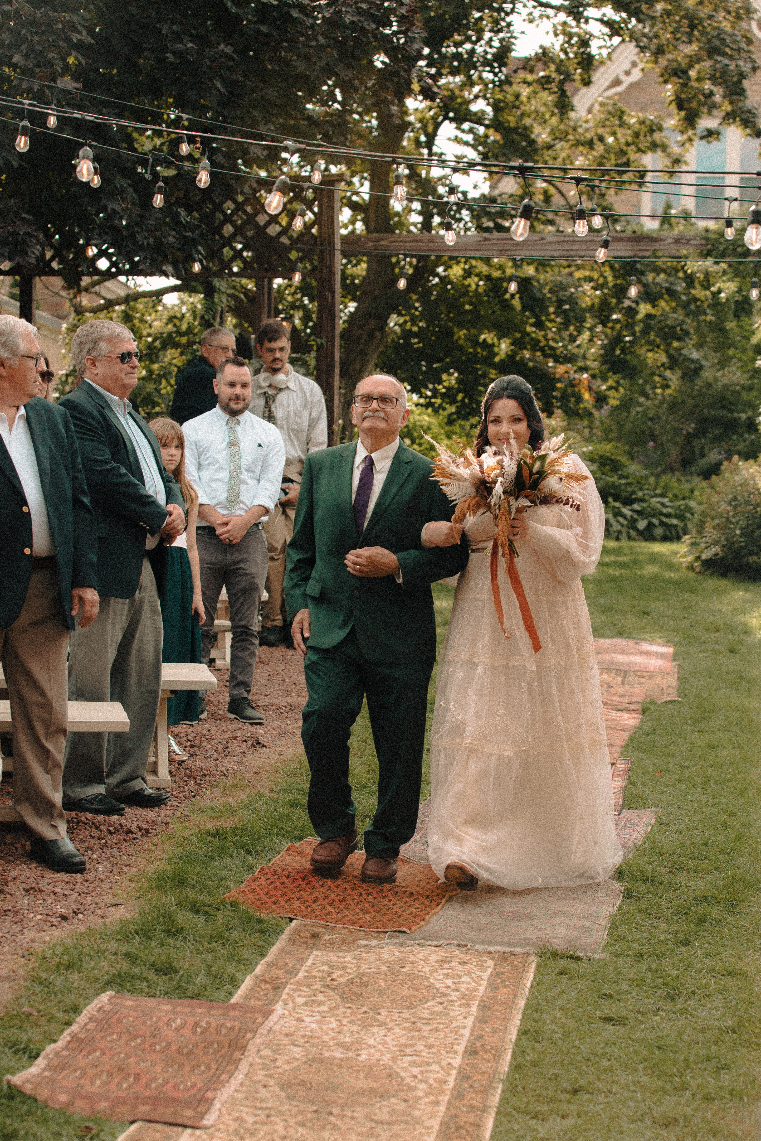 Bride and her father walking down a ceremony aisle made up of vintage rugs under twinkle lights in a garden
