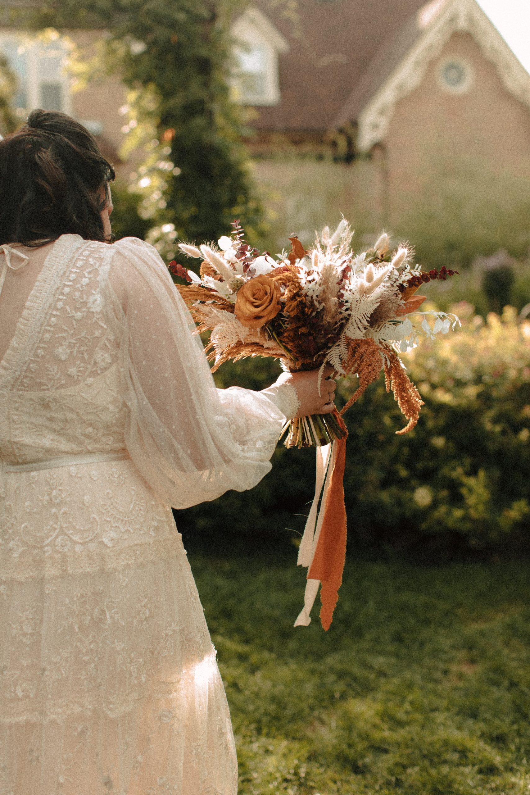 Bride with boho lace dress carrying her red and orange flowers in the sunlight of a garden