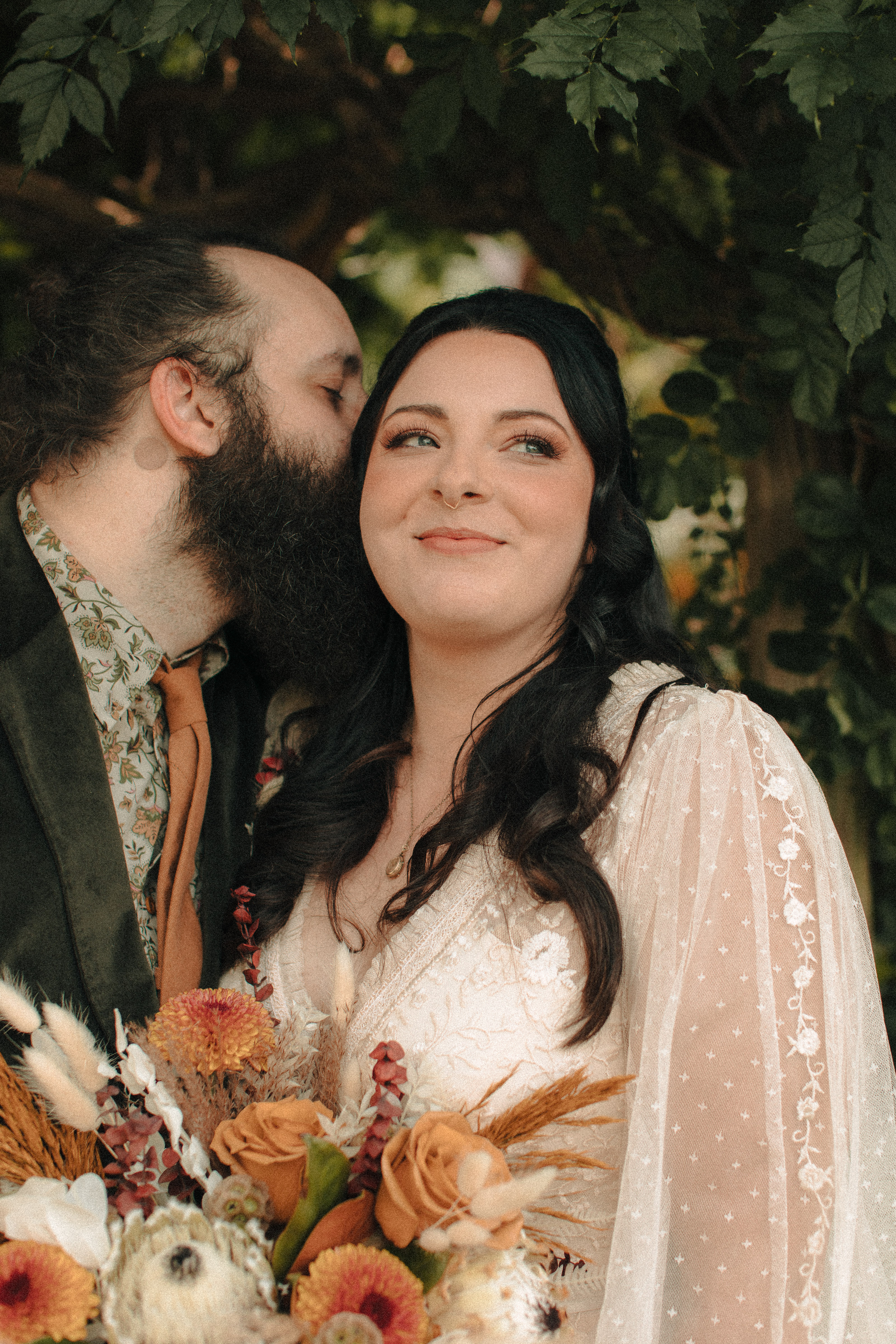 Groom with a beard and man bun kissing his new wife's cheek, as the wife smiles into the distance with Priscilla Presley inspired hair and Makeup