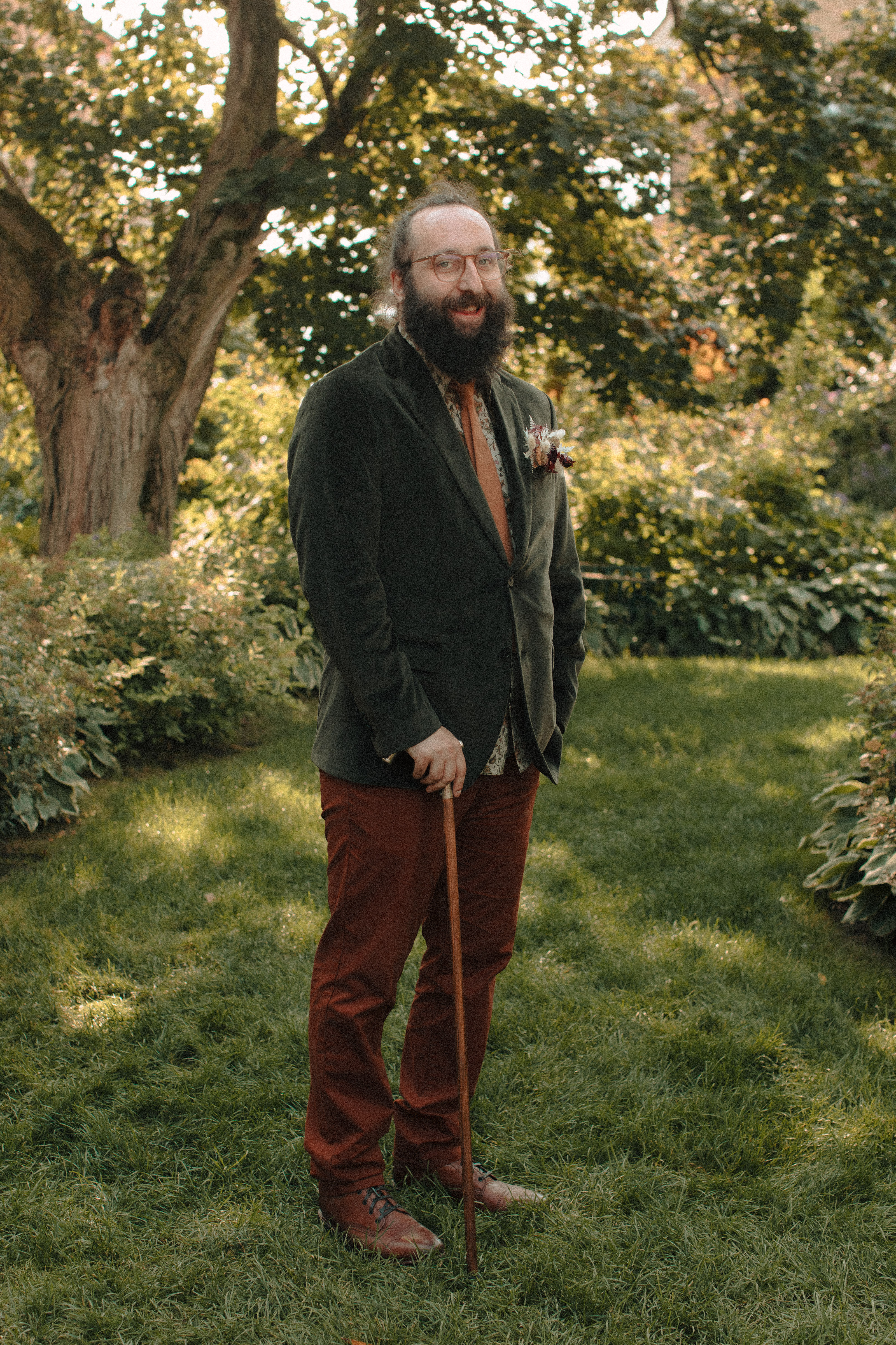 Groom in green velvet coat, tan pants, and caramel tie standing in a garden