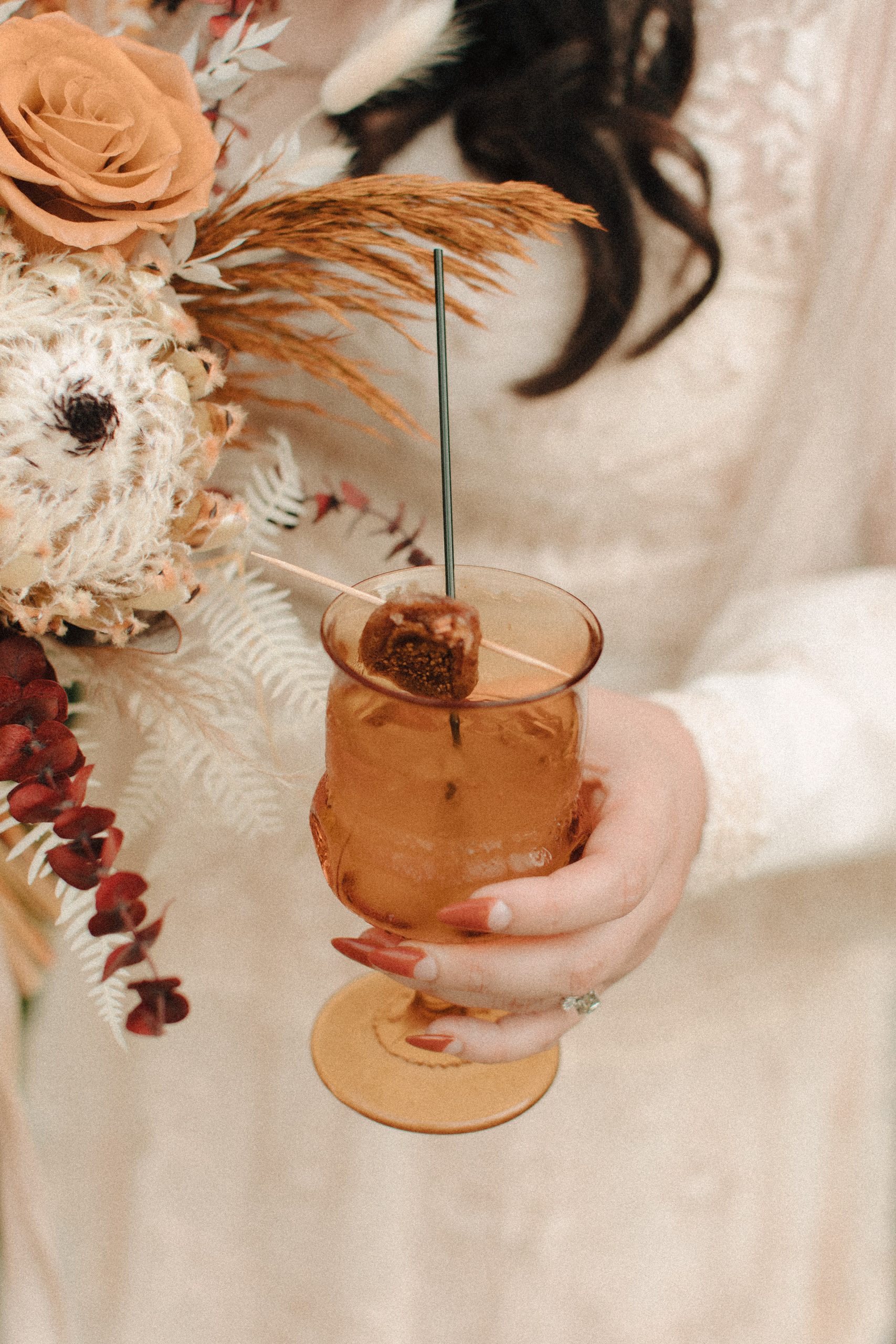 Fancy wedding cocktail in a bride's hand with her flowers in the background. Drink is a fig jam infused gin cocktail