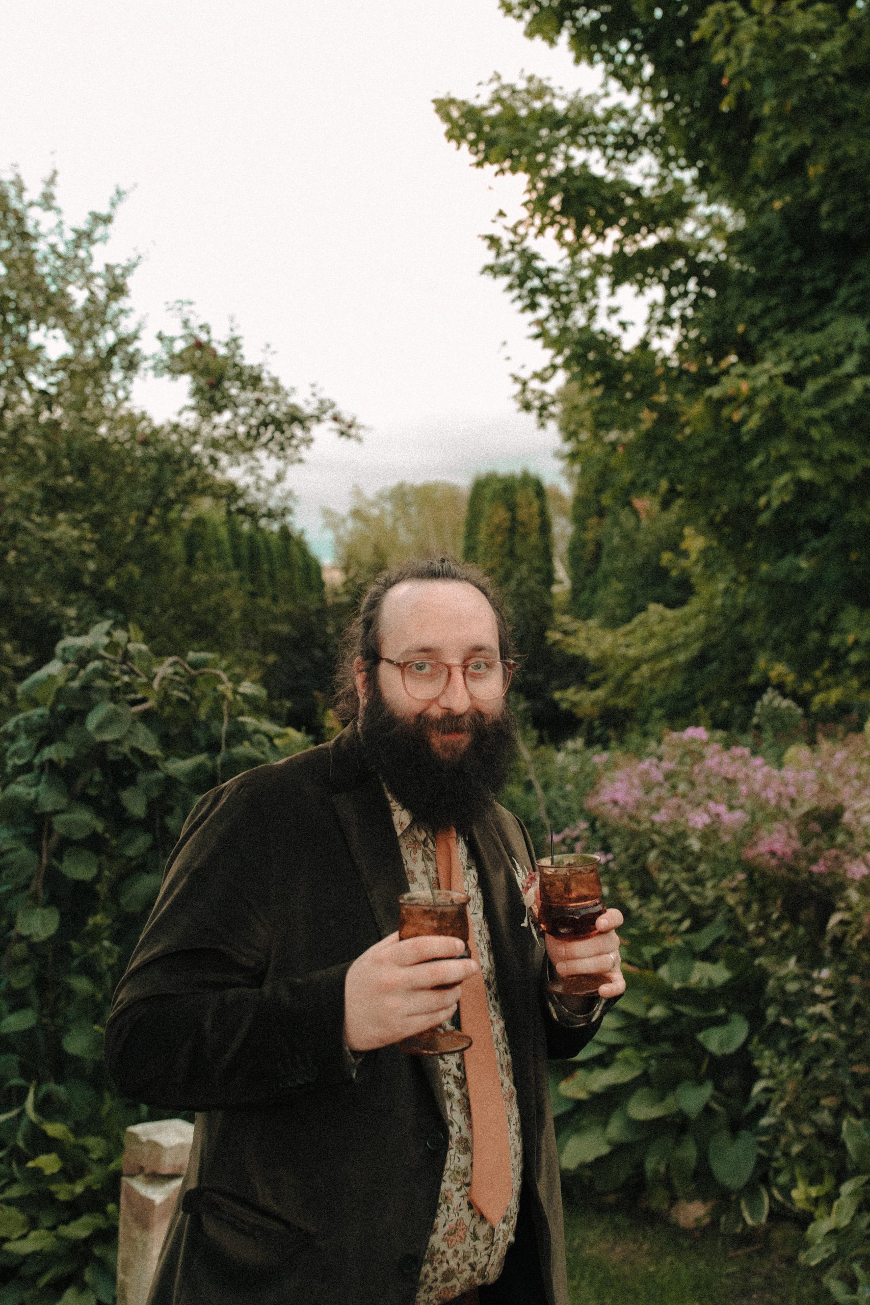 Groom holding two amber cocktail glasses in a garden