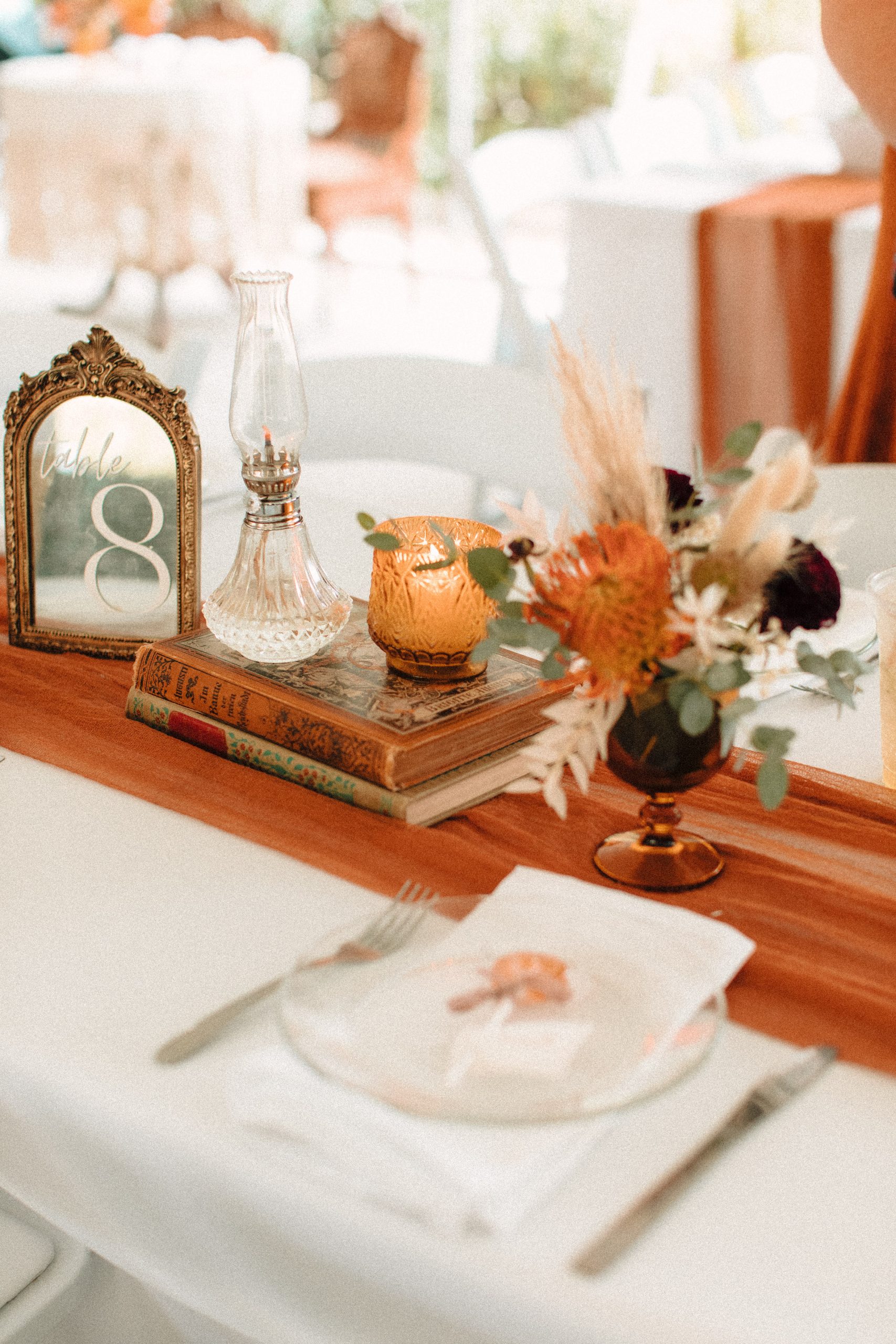 Victorian inspired wedding table centerpeces with orange runners, old books, candles, and mirrored table numbers with florals in a goblet