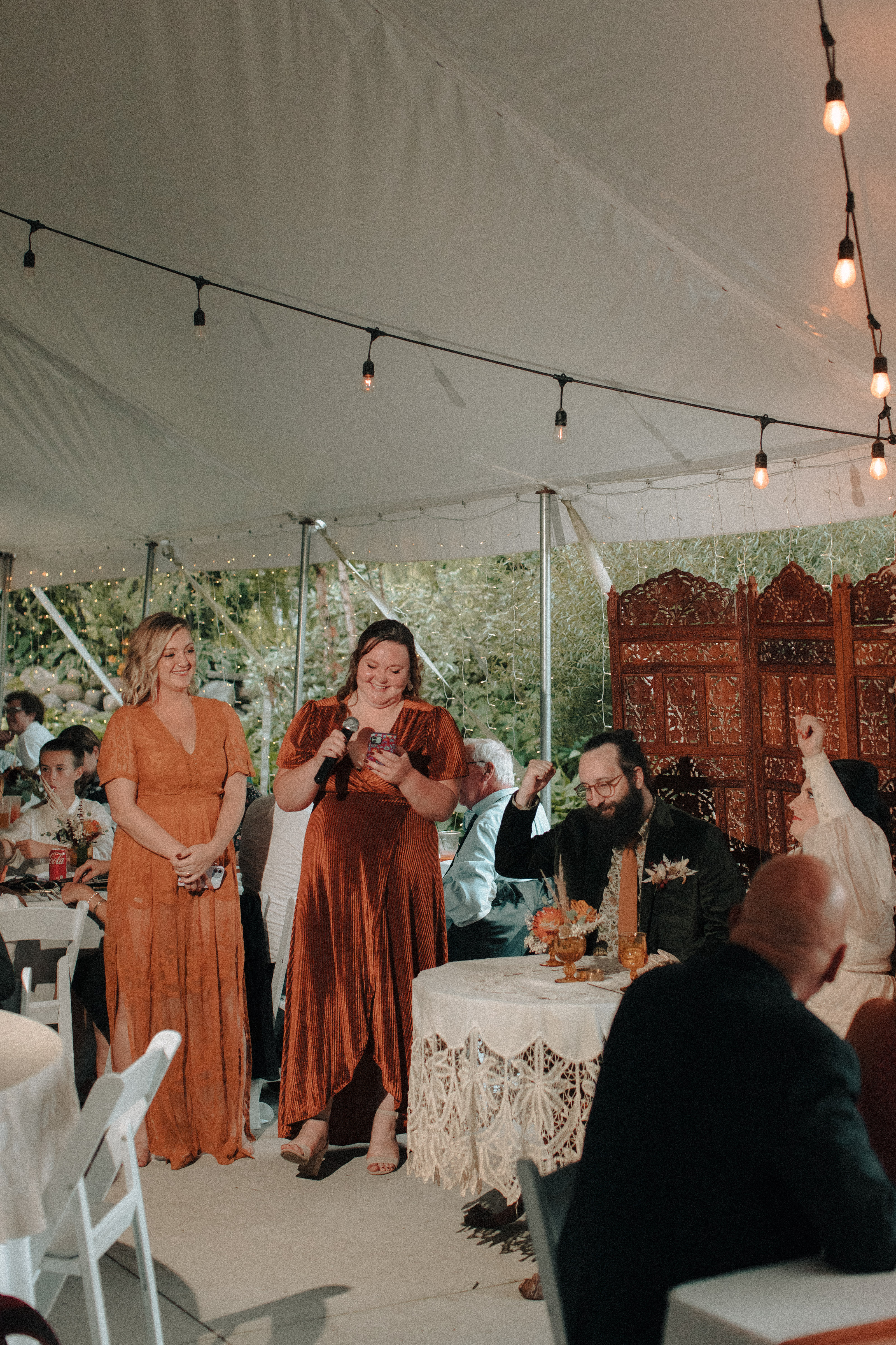Bridesmaids in orange dresses giving a combined wedding speech as bride and groom cheer them on